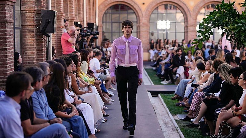 Desfile de moda con modelo masculino en pasarela, arquitectura de ladrillo con arcos y el número "1886" al fondo. Audiencia presente.