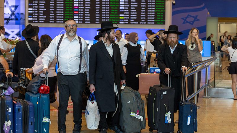 Terminal del Aeropuerto Ben Gurion en Tel Aviv, Israel, con viajeros, incluyendo hombres Haredi, equipaje y la bandera de Israel visible.