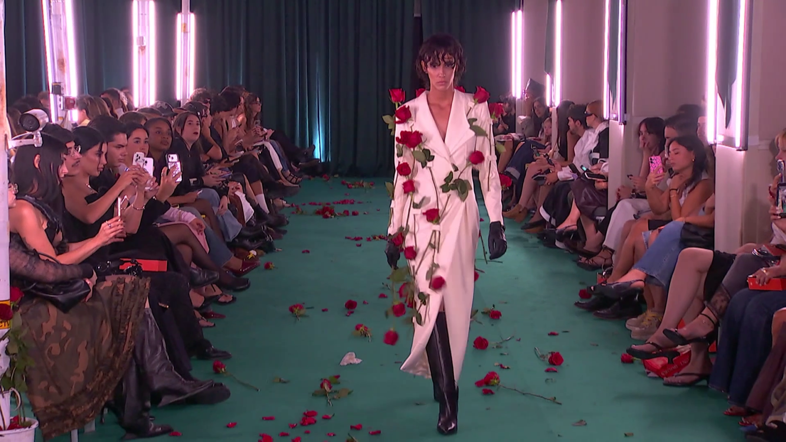 Modelo con abrigo blanco y rosas rojas desfila por pasarela floral, público graba con móviles, iluminación rosada. Desfile Calafat, Mercedes-Benz Fashion Week Madrid.