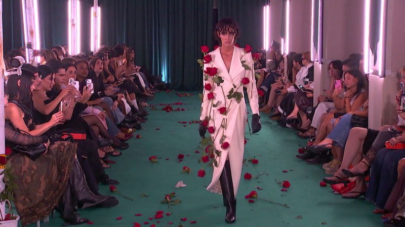 Modelo con abrigo blanco y rosas rojas desfila por pasarela floral, público graba con móviles, iluminación rosada. Desfile Calafat, Mercedes-Benz Fashion Week Madrid.