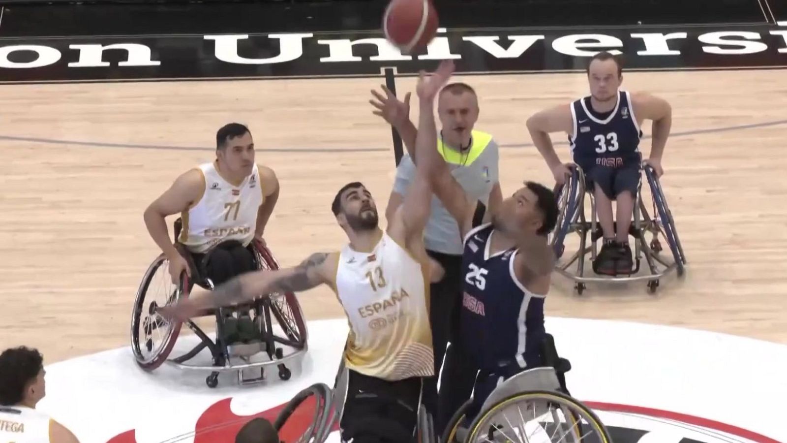 Jugadores de baloncesto en silla de ruedas compiten por el balón en el aire. Se aprecian camisetas con "ESPAÑA", números "13" y "25", y un árbitro observando.