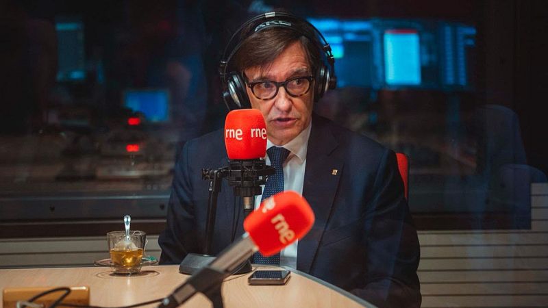 Primer plano de Salvador Illa en un estudio de radio, con auriculares y frente a micrófonos de RNE. Se observa una mesa con un vaso y un móvil.