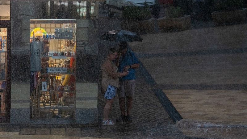 Dos personas comparten un paraguas bajo lluvia intensa en una calle urbana, observando un móvil. A la izquierda, el escaparate de una tienda con el cartel "CARTAGENA".