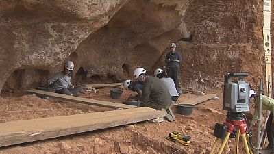 Arque�logos en excavaci�n con cascos blancos, trabajando en tierra rojiza. Se aprecian marcadores estratigr�ficos "GII" y "GI" en la pared, junto a equipo de topograf�a y tablones de madera.
