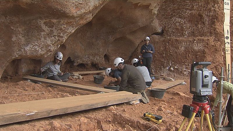 Arqueólogos en excavación con cascos blancos, trabajando en tierra rojiza. Se aprecian marcadores estratigráficos "GII" y "GI" en la pared, junto a equipo de topografía y tablones de madera.