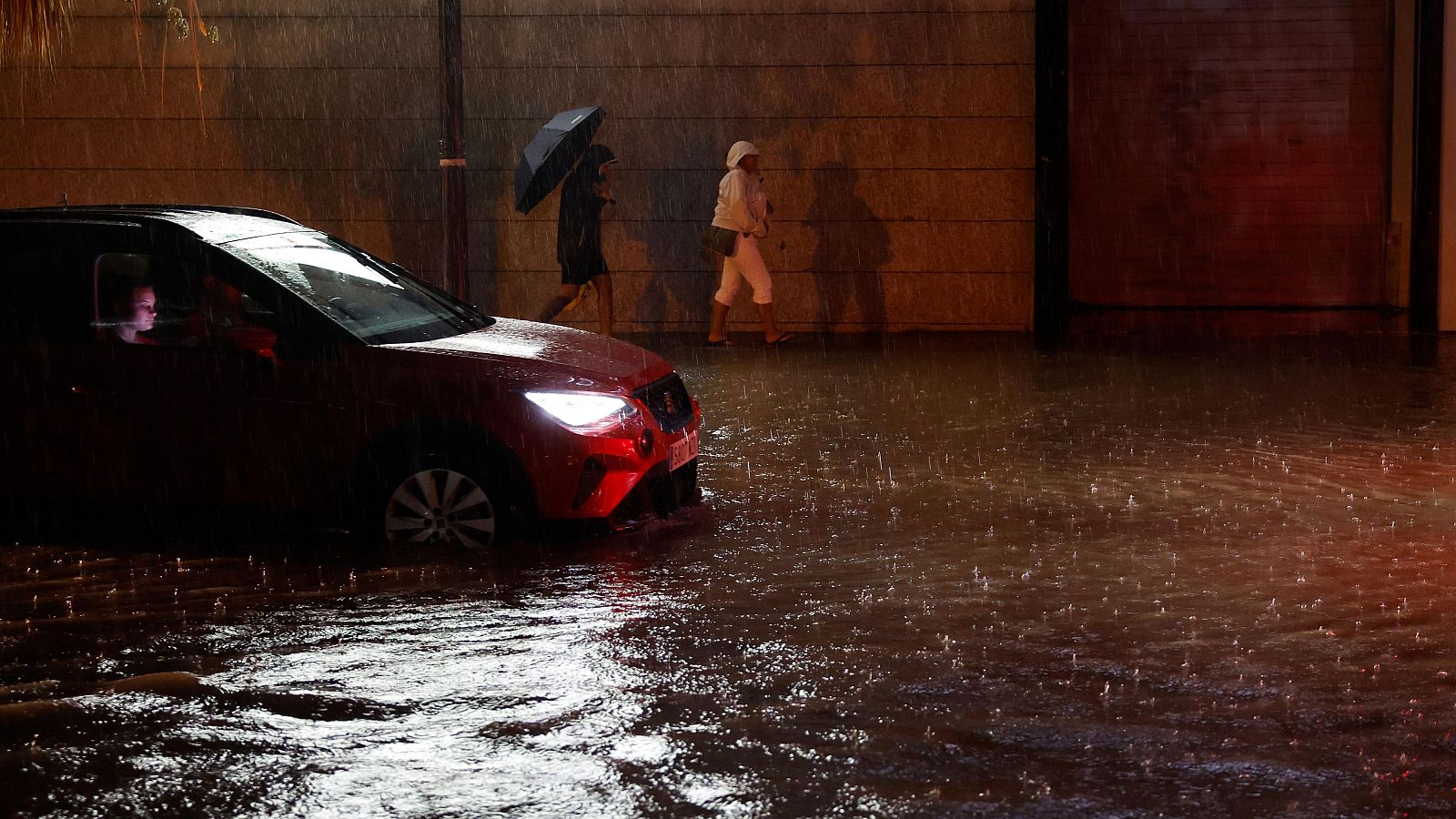 Noche de fuerte lluvia e inundación en la Comunidad Valenciana. Un coche rojo oscuro con faros encendidos y dos personas sorteando el agua en una calle oscura con pared de ladrillo al fondo.