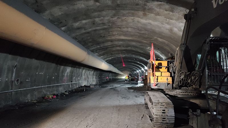 Interior de túnel en construcción con excavadora VOLVO, trabajadores de seguridad, tubería y punto láser rojo. Obras de ingeniería civil.