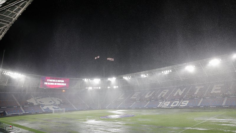 Estadio de fútbol de noche con lluvia intensa. Pantalla LED anuncia suspensión del partido. Se lee "LEVANTE 1909" en las gradas. Campo mojado con charcos.