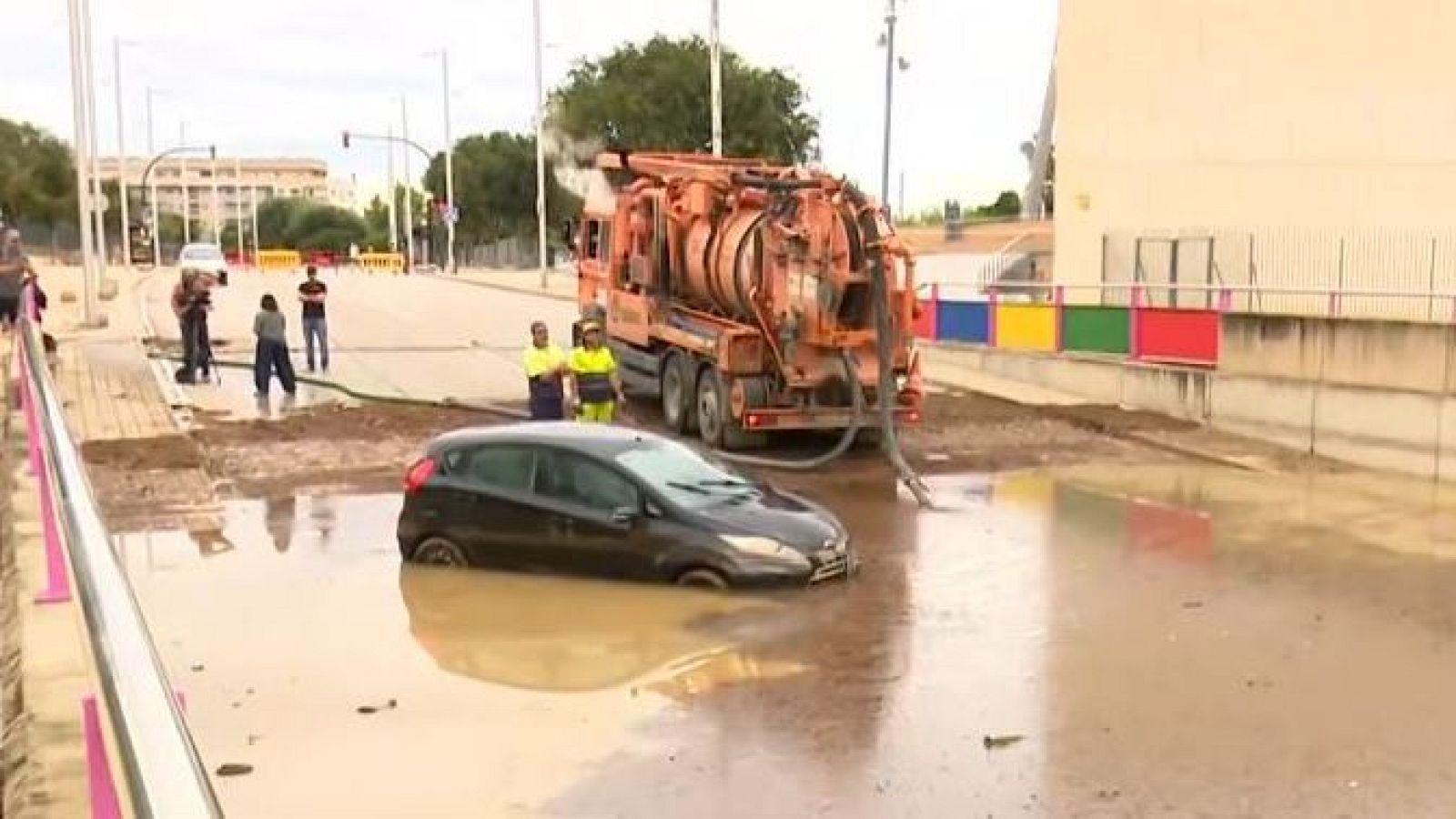 Coche atrapado en inundación. Camión de succión naranja drenando agua. Trabajadores y equipo de medios presentes.