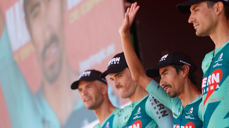 Ciclistas del equipo Kern Pharma saludan con maillots verde azulado y gorras negras. Fondo con logos "KERN PHARMA", "Telenavarra" y rostro desenfocado.