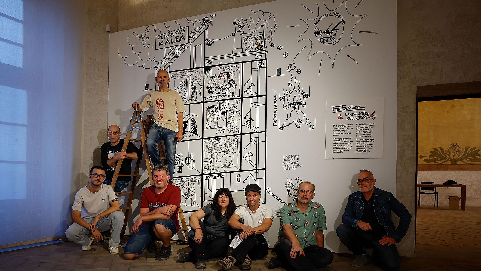Grupo de personas posando frente a un mural de cómic en blanco y negro con el título "Faltabezes & Kanpai Jotze & Kolektiboa".