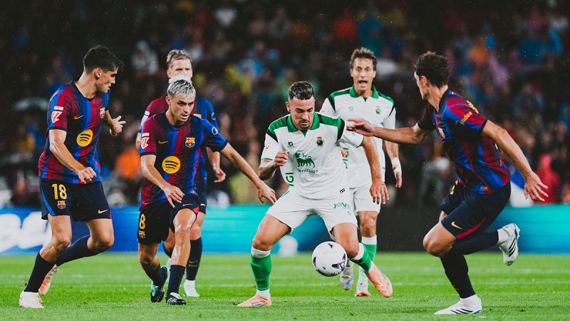Arana del Racing de Santander disputa el balón contra jugadores del FC Barcelona en un partido. Se aprecian los escudos y patrocinadores de ambos equipos.