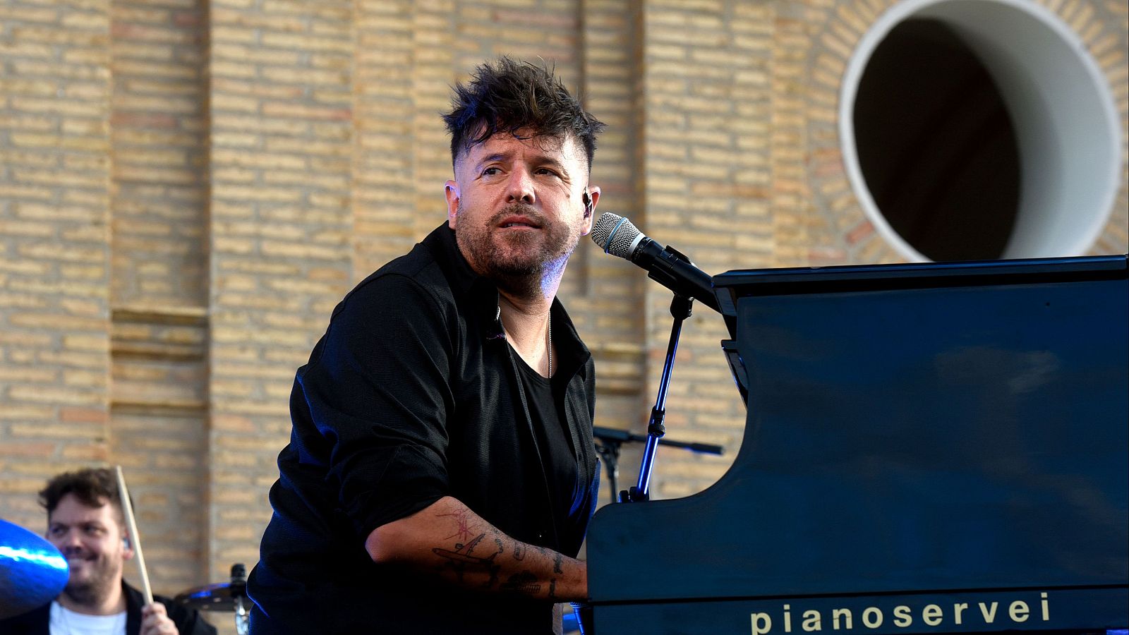 Pablo López, cantante y pianista, actúa frente a un piano azul oscuro. Viste de negro, con barba y tatuajes. A su lado, un batería sonríe. El fondo es una pared de ladrillo con un óculo.