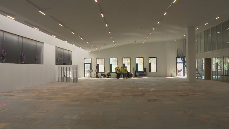 Interior de edificio en construcción con personal de obra y equipos de medición. Se aprecian ventanales, una estructura de dos niveles y una columna blanca.