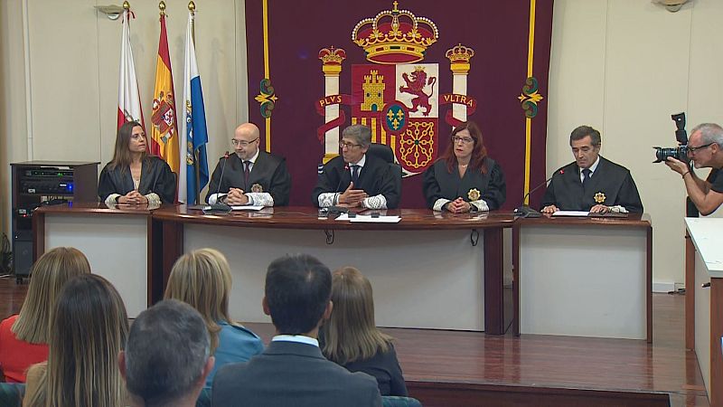 Cinco funcionarios judiciales con togas negras participan en un acto solemne, con banderas y un escudo al fondo. Un camarógrafo documenta la ceremonia.