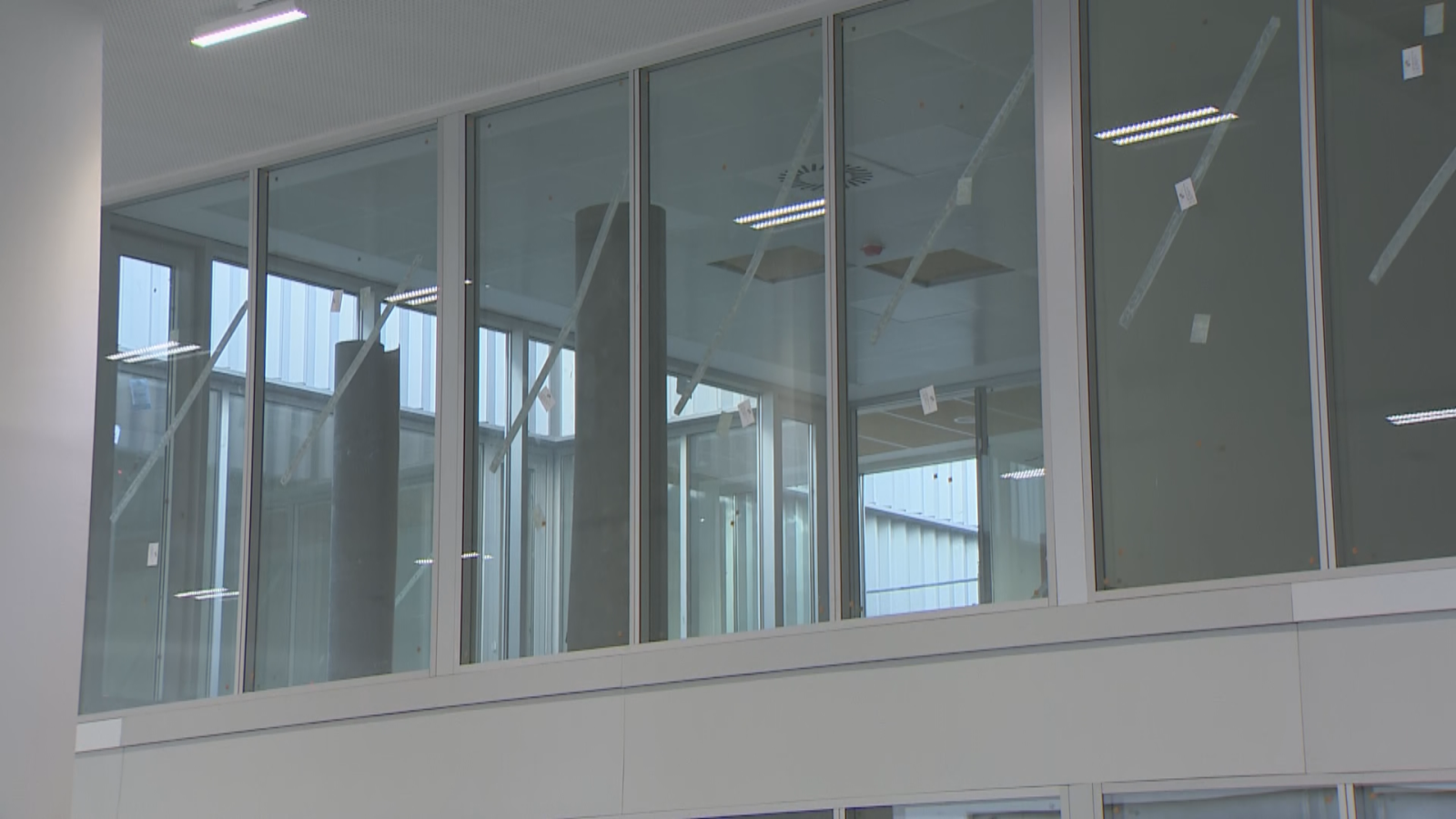 Interior de edificio moderno con amplios ventanales que revelan un espacio en construcción o industrial. Se ven vigas, chapa ondulada y pegatinas blancas en los cristales.