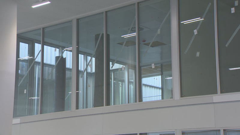 Interior de edificio moderno con amplios ventanales que revelan un espacio en construcción o industrial. Se ven vigas, chapa ondulada y pegatinas blancas en los cristales.