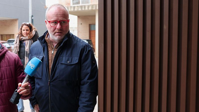 Ximo Puig, con gafas rojas y chaqueta azul, es entrevistado por TVE en un entorno urbano con edificios y una fachada moderna de listones verticales.