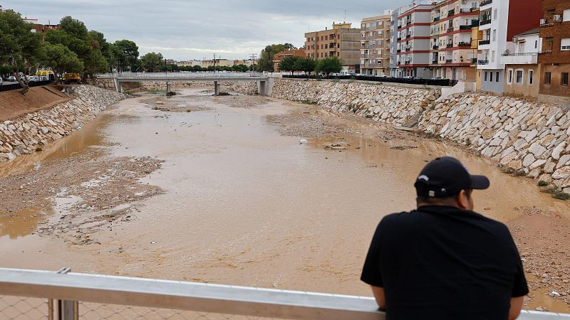 Vista panorámica de un cauce de agua bajo un cielo nublado, con un individuo observando el bajo nivel y turbidez del agua. Edificios residenciales y vegetación flanquean el cauce, con una excavadora visible.