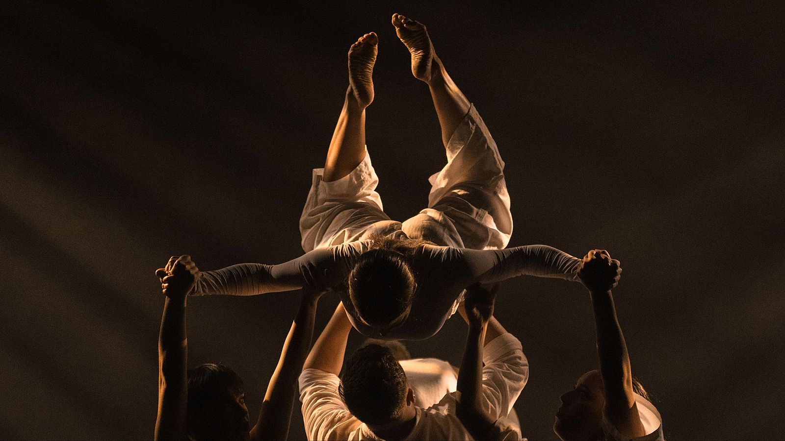 Artistas en un escenario oscuro realizan una compleja figura de equilibrio y fuerza bajo un potente haz de luz, destacando la colaboraci�n y destreza f�sica.