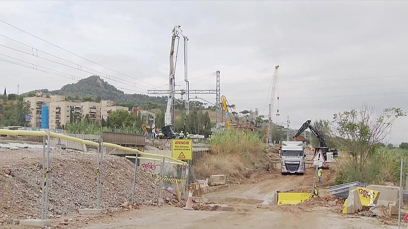 Obra de construcción con bomba de hormigón, camión DAF y cartel "ATENCIO: Pas restringit vehicles d'obra" bajo cielo nublado. Se aprecian edificios y montaña al fondo.