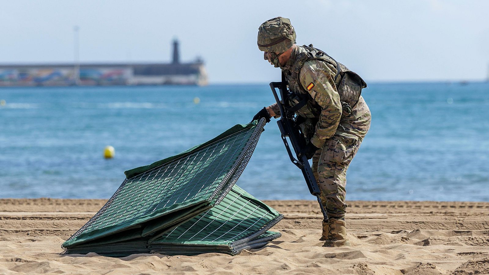 Soldado español en playa con uniforme pixelado, casco y chaleco táctico, manipulando paneles de malla metálica sobre arena. Mar azul y boya amarilla al fondo.