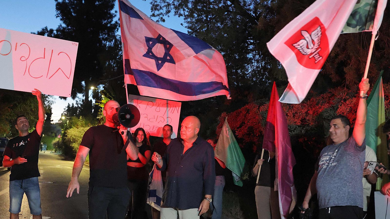Manifestación nocturna en Israel con pancartas en hebreo, la bandera israelí y un hombre arengando a la multitud con un megáfono.