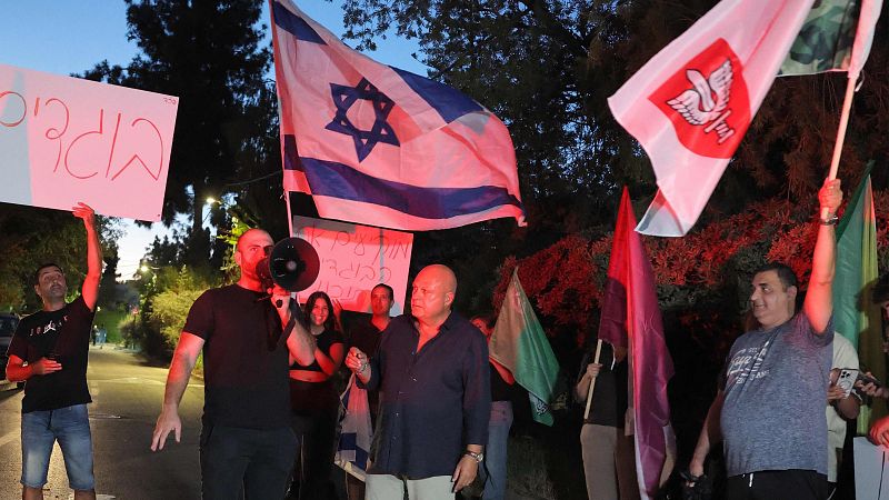 Manifestación nocturna en Israel con pancartas en hebreo, la bandera israelí y un hombre arengando a la multitud con un megáfono.