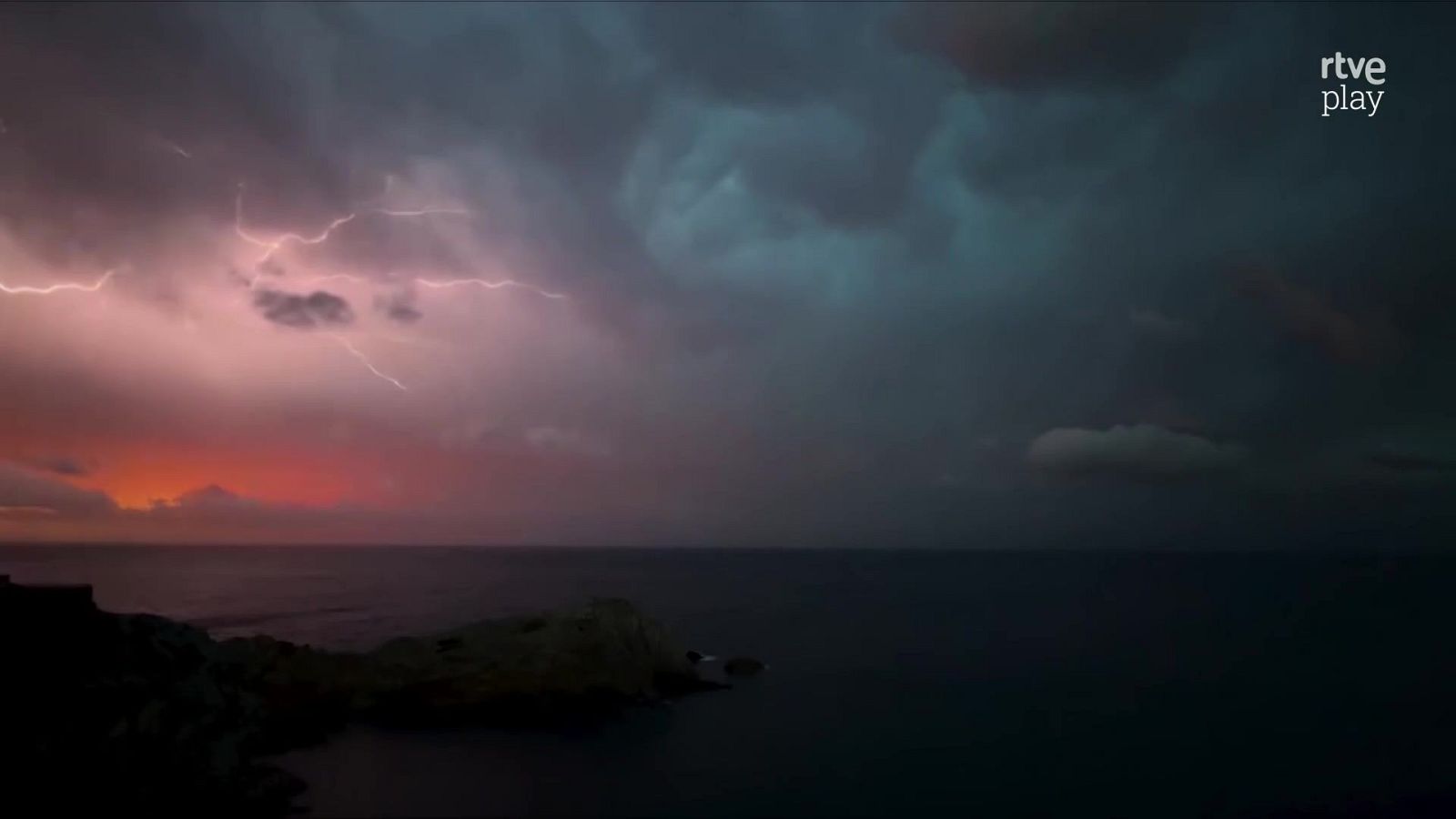 Cielo tormentoso con relámpagos y nubes de colores cálidos y fríos sobre un mar oscuro y una costa rocosa. Logo de "rtve play" en la esquina superior derecha.
