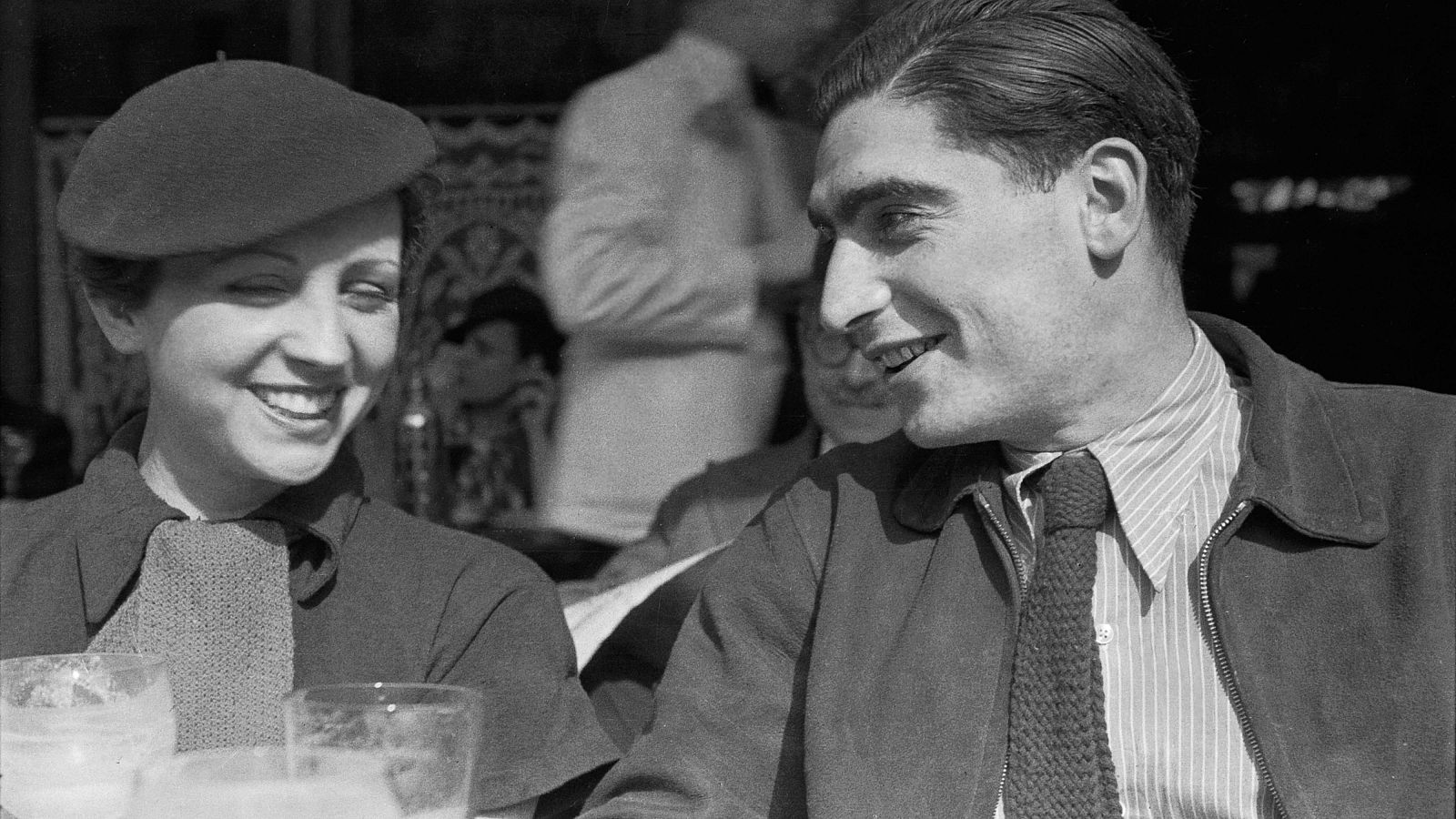 Gerda Taro y Robert Capa, fotógrafos, sonríen sentados en una terraza de café, con vasos en primer plano. Fotografía histórica en blanco y negro.
