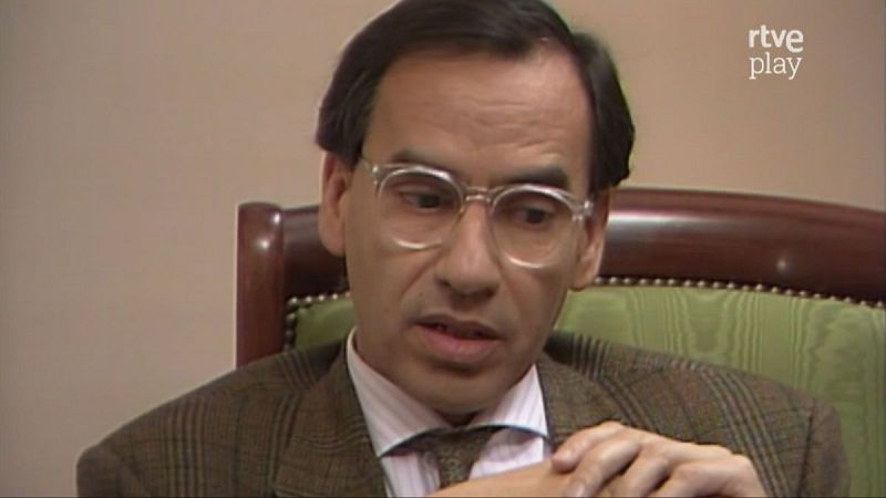 Primer plano de Alfonso Guerra con traje a cuadros, camisa de rayas y gafas gruesas. Fondo liso con logo de "rtve play".