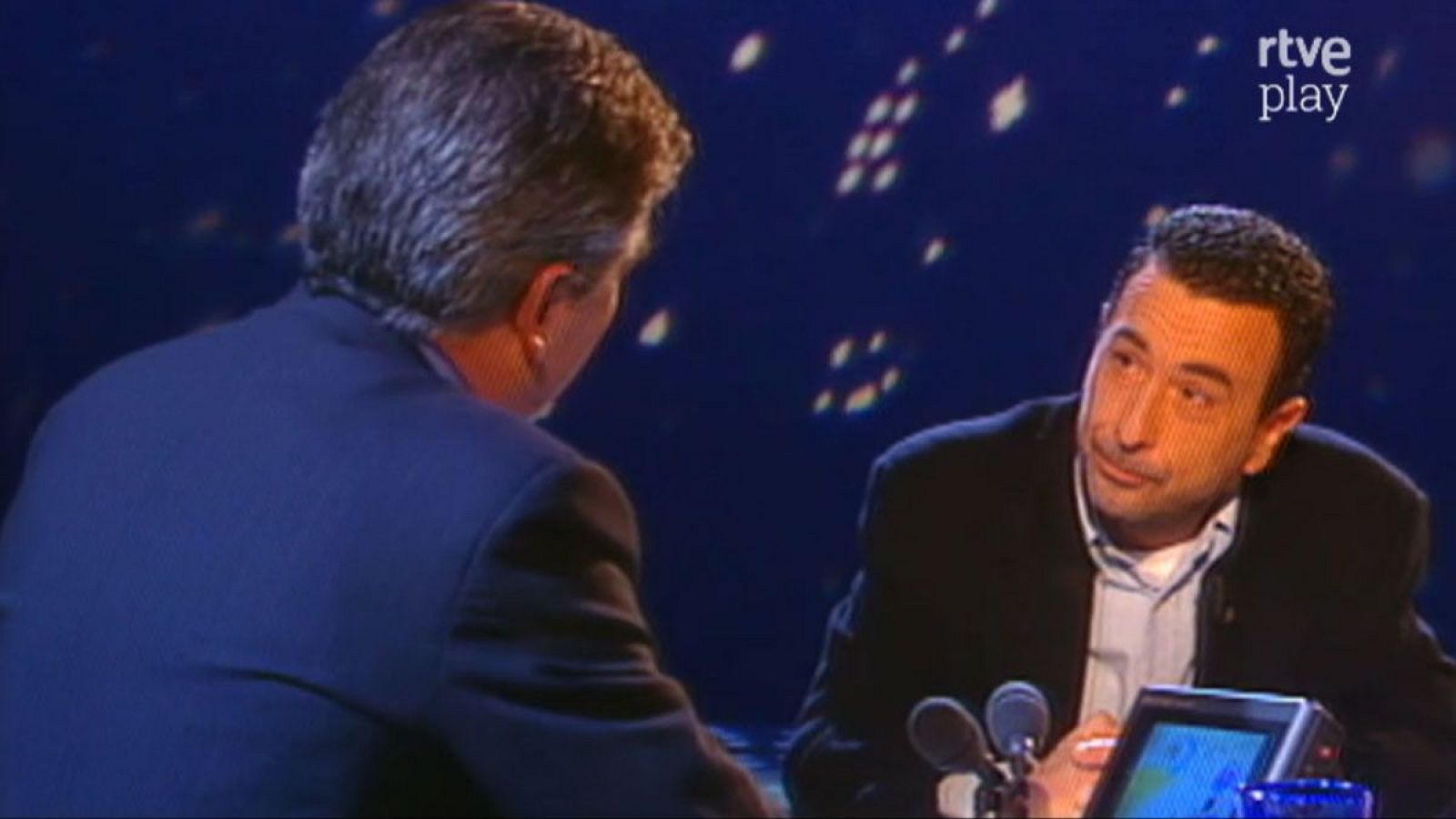 Pedro Ruiz y Jose Luis Garci en un plató de "rtve play". Ruiz, de frente, con manos entrelazadas y micrófonos delante. Garci, de espaldas, de perfil, con cabello canoso.