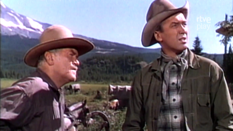 James Stewart y Walter Brennan contemplan un paisaje montañoso, con Stewart a la derecha y Brennan a su izquierda, ambos con sombreros de vaquero.