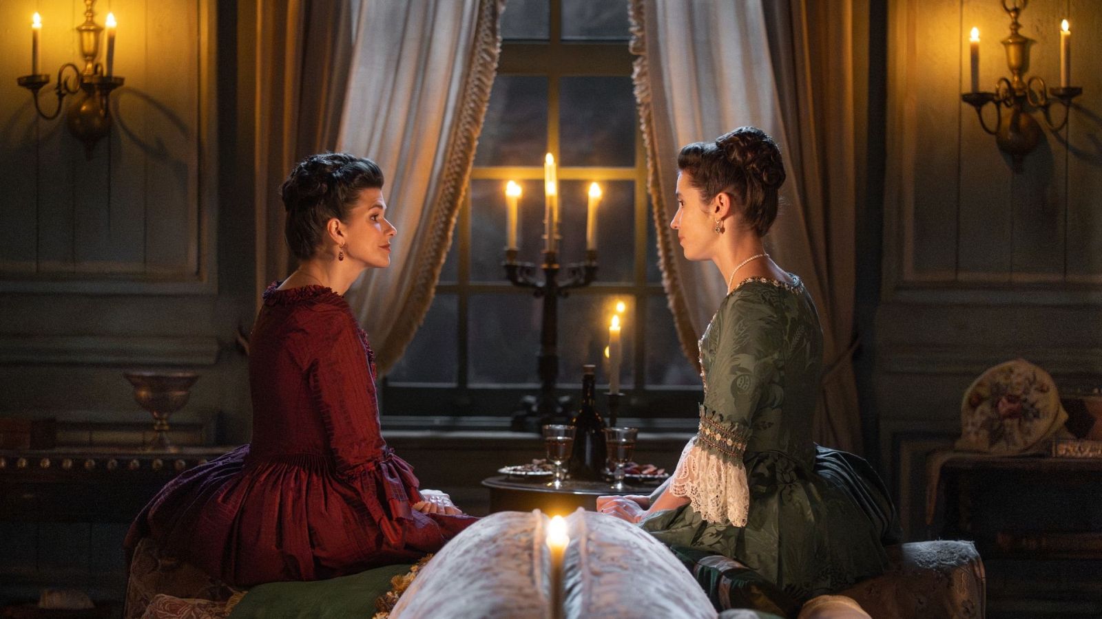 Dos actrices vestidas de época, comparten un momento íntimo a la luz de las velas en una escena histórica.
