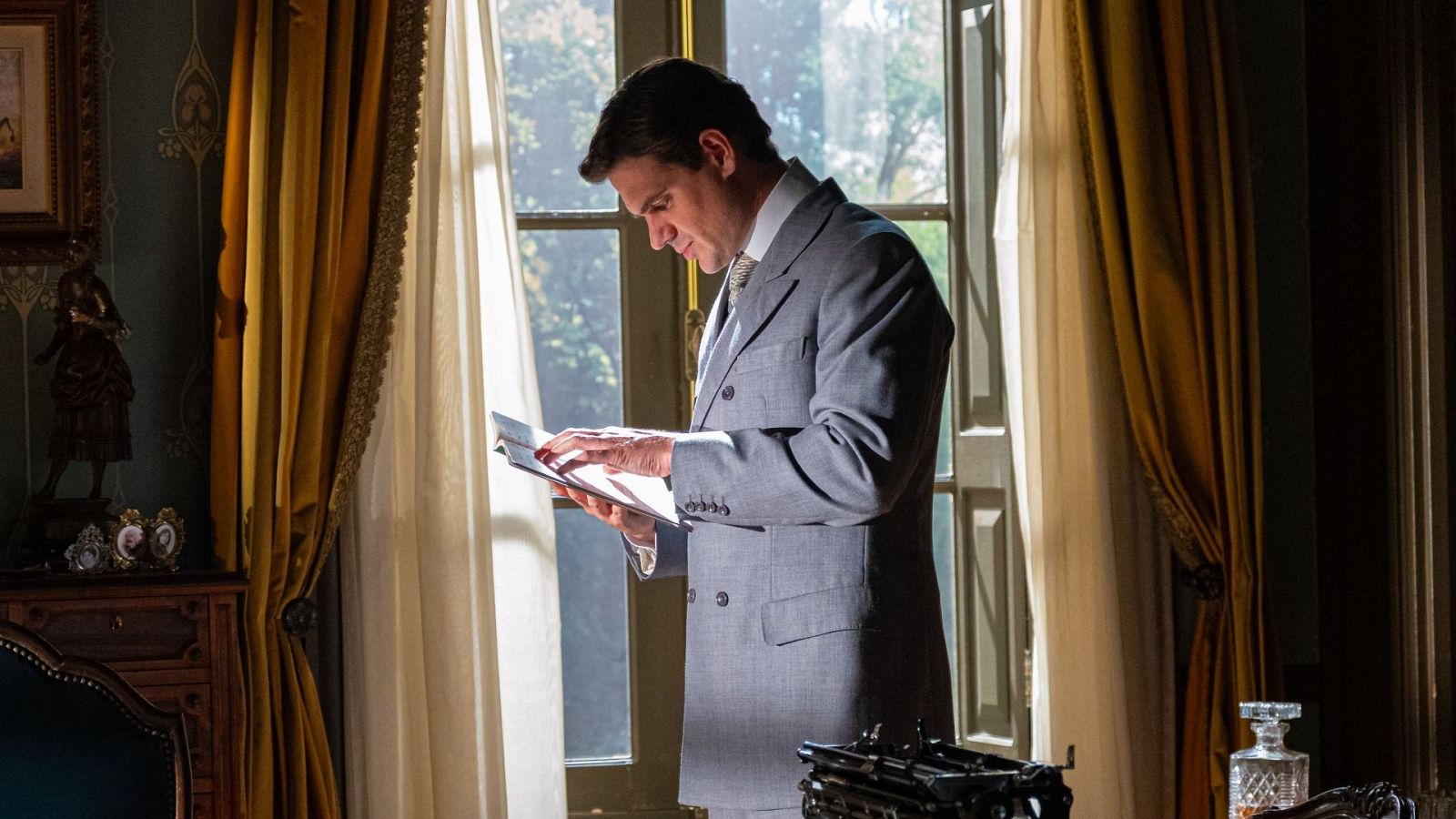 Hombre con traje gris y corbata estampada lee un documento en una estancia clásica con gran ventana y decoración opulenta. Máquina de escribir antigua y licorera en primer plano.