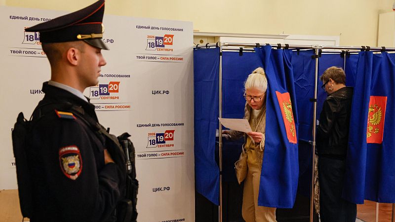 Escena de votación en Rusia con un agente de seguridad y ciudadanos en cabinas. Se aprecian emblemas del águila bicéfala y textos en cirílico como 'Día Único de Votación' y 'Tu Voto – La Fuerza de Rusia'.