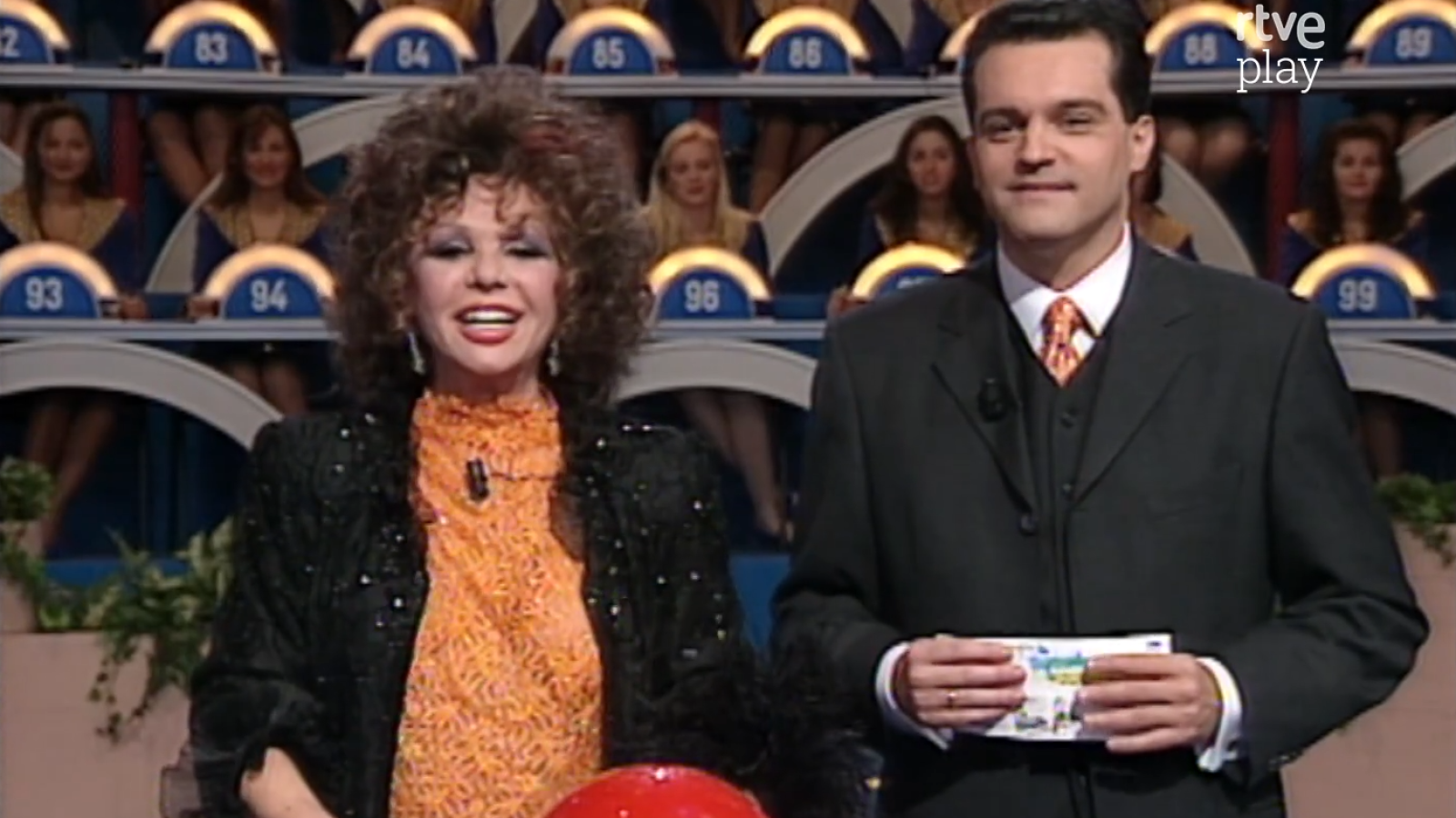 Marujita Díaz y Jordi Hurtado en un plató de RTVE con concursantes en cabinas numeradas y el logo "tve play".