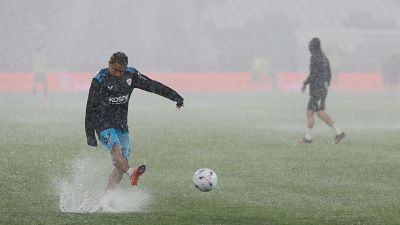 Jugador de f�tbol con trenzas y gafas patea un bal�n bajo intensa lluvia, salpicando agua. Viste camiseta oscura con logo "KOSNE" y pantalones azul claro.