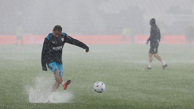 Jugador de fútbol con trenzas y gafas patea un balón bajo intensa lluvia, salpicando agua. Viste camiseta oscura con logo "KOSNE" y pantalones azul claro.