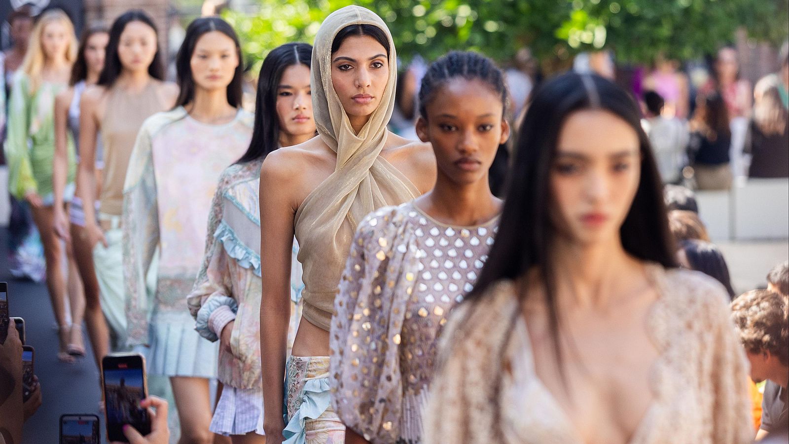 Modelos desfilan en pasarela exterior en la Mercedes-Benz Fashion Week Madrid, luciendo prendas de Custo Barcelona con brillos y colores pastel. Público graba el evento con móviles.