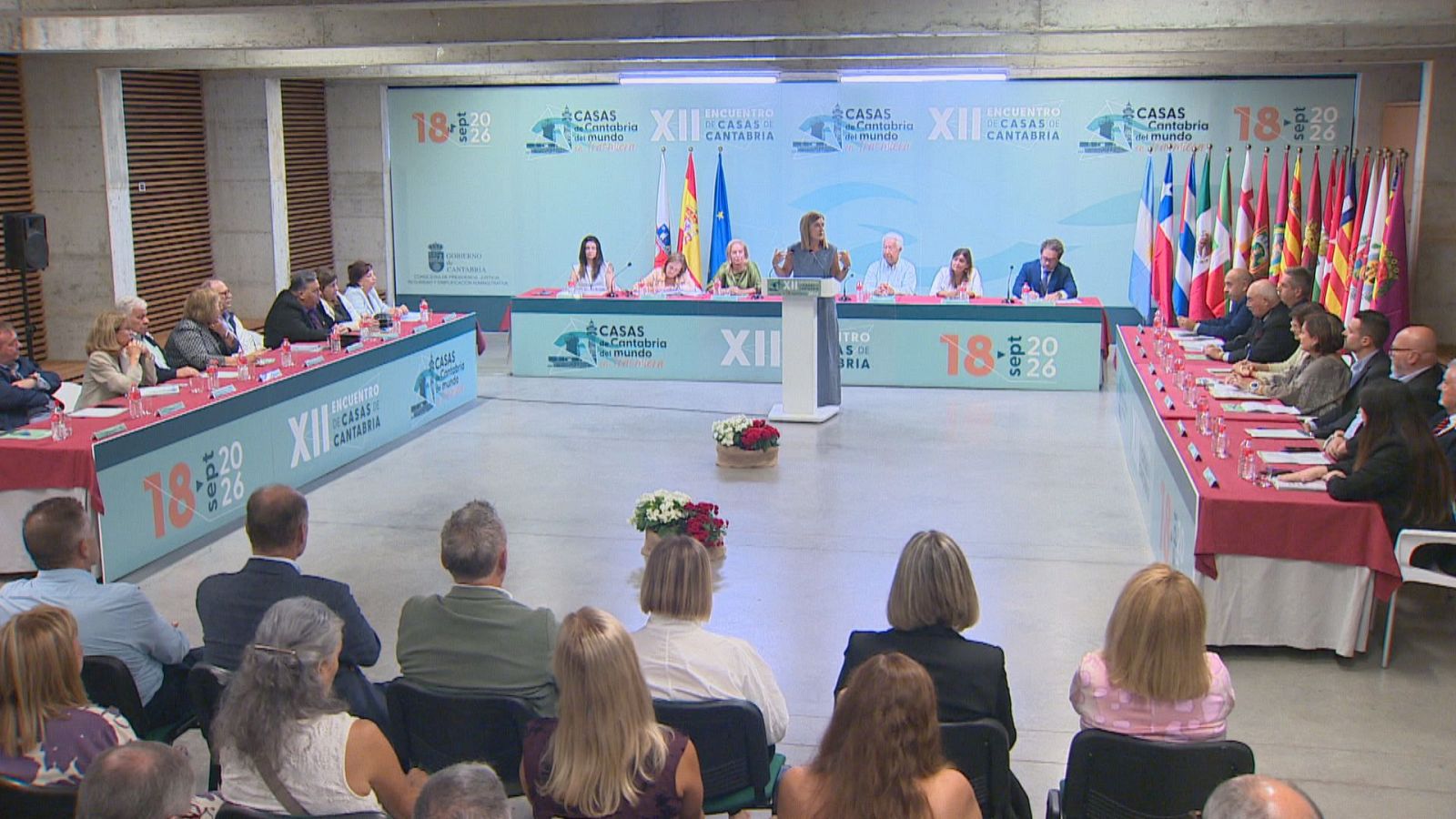 María José Sáenz de Buruaga habla en el XII Encuentro de Casas de Cantabria, con la mesa presidencial y banderas de España y Cantabria de fondo.