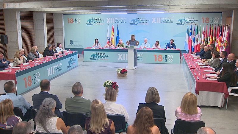 María José Sáenz de Buruaga habla en el XII Encuentro de Casas de Cantabria, con la mesa presidencial y banderas de España y Cantabria de fondo.