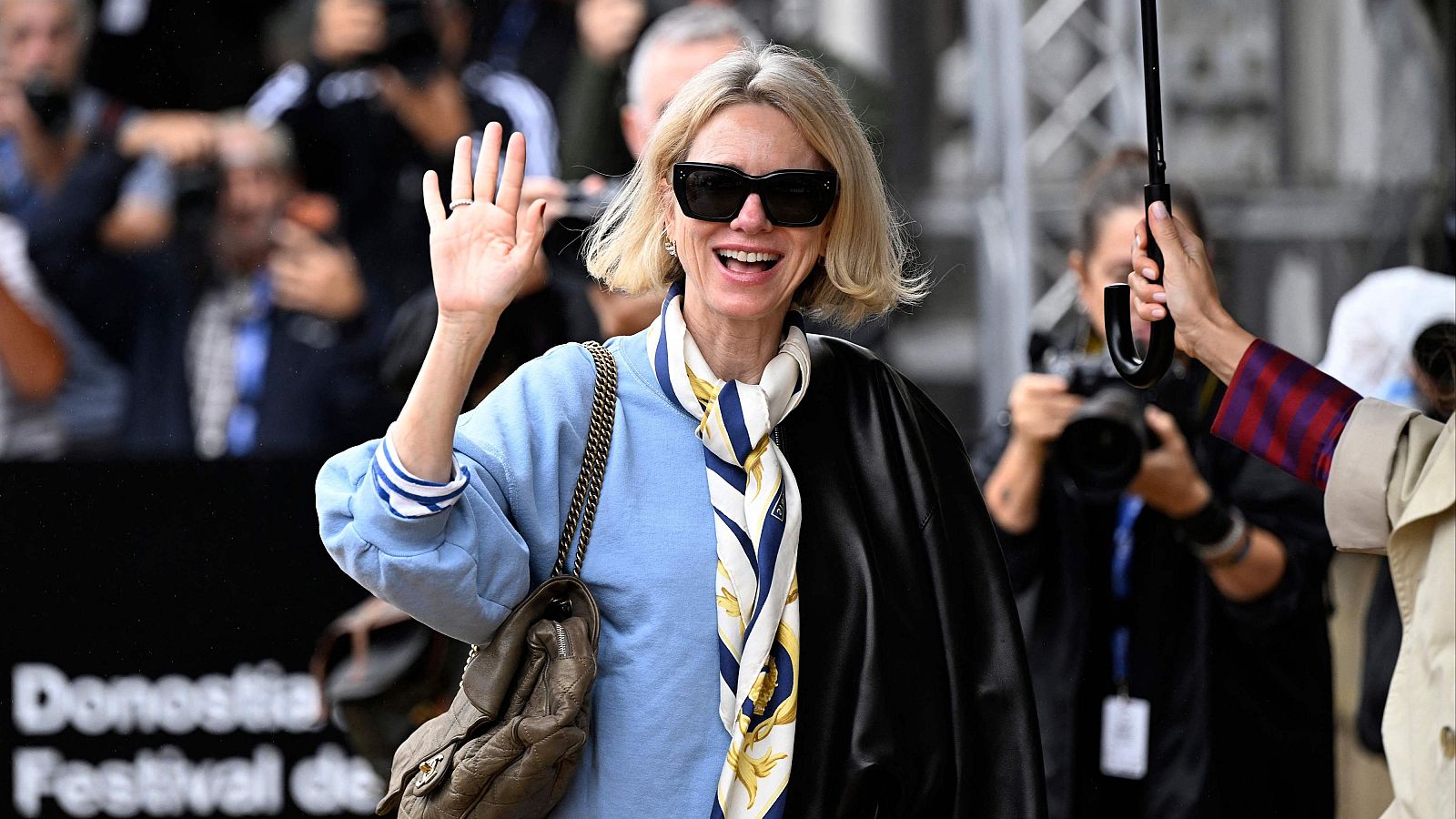 Naomi Watts saluda sonriente en el Festival de Cine de San Sebastián, luciendo gafas de sol, pañuelo de seda y blusa azul. Se aprecian fotógrafos y un cartel con "Donostia Festival de".