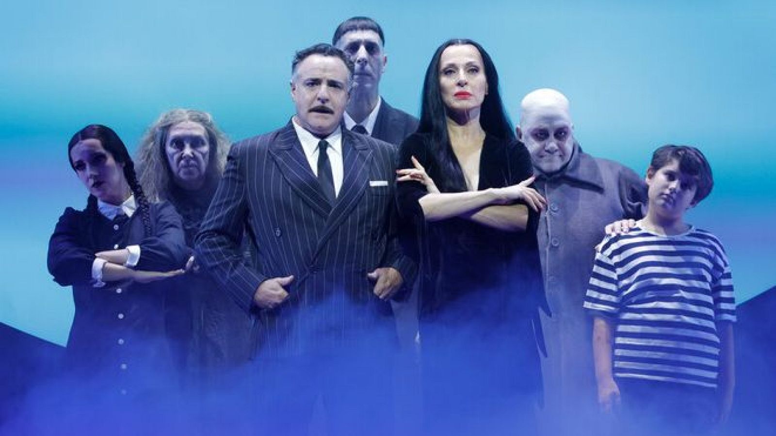 Actores de 'La Familia Addams' posan en un escenario teatral con fondo azul y humo. Se identifican Gómez (Xavi Mira), Morticia (Lydia Fairén), Miércoles (Carla Postigo), Tío Fétido (David Ávila), Abuela (Meritxell Duró) y Largo (Javier Canales).