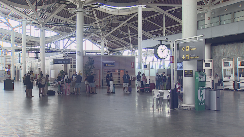 Terminal de aeropuerto con pasajeros, señales de "Llegadas" y "Facturación", un reloj marcando las 12:10, un AED/DEA y quioscos numerados.