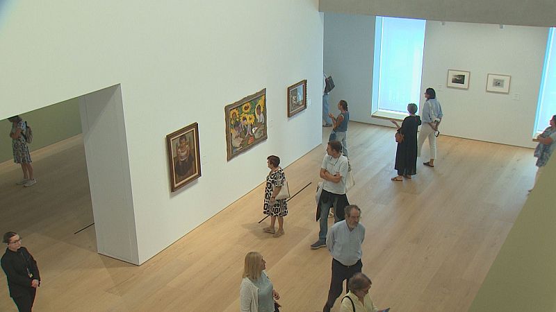 Galería de arte moderna con obras expuestas y visitantes observando. Se aprecian cuadros de diferentes tamaños y estilos, incluyendo una escena con girasoles.