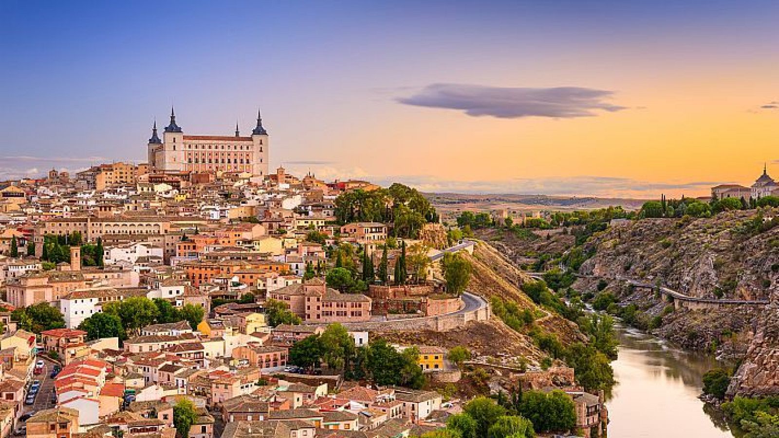 Vista panorámica de Toledo, con el Alcázar y la ciudad sobre una colina, flanqueada por el río Tajo en un barranco rocoso, bajo un cielo de atardecer/amanecer.