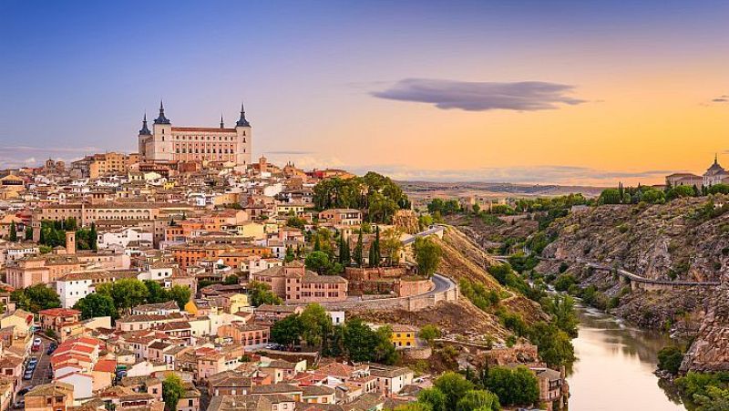 Vista panorámica de Toledo, con el Alcázar y la ciudad sobre una colina, flanqueada por el río Tajo en un barranco rocoso, bajo un cielo de atardecer/amanecer.