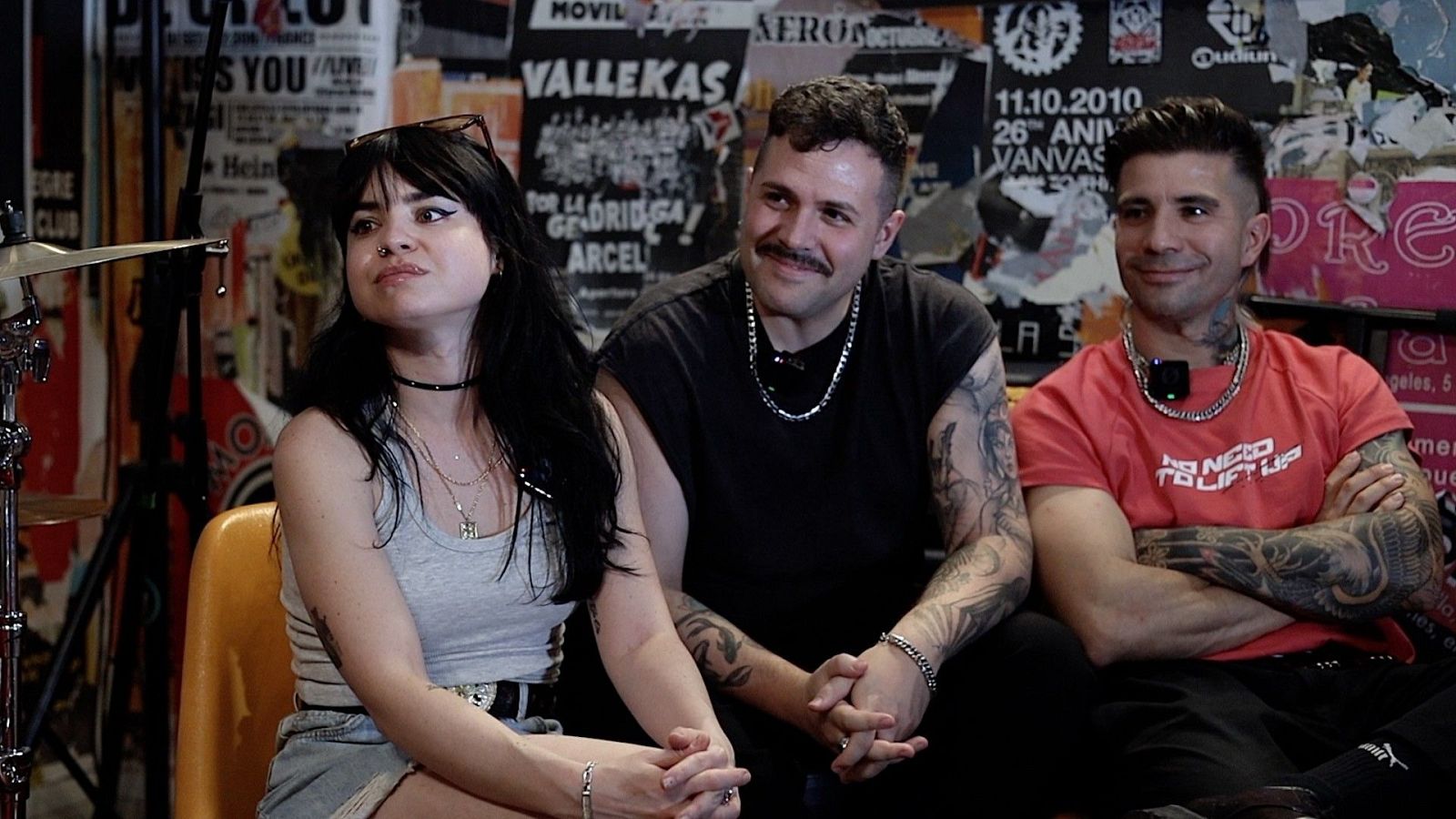 Omar Montes, El Bobe y una mujer posan sonrientes en un local con pared de carteles. Se aprecian tatuajes, cadenas y un set de bater�a al fondo.