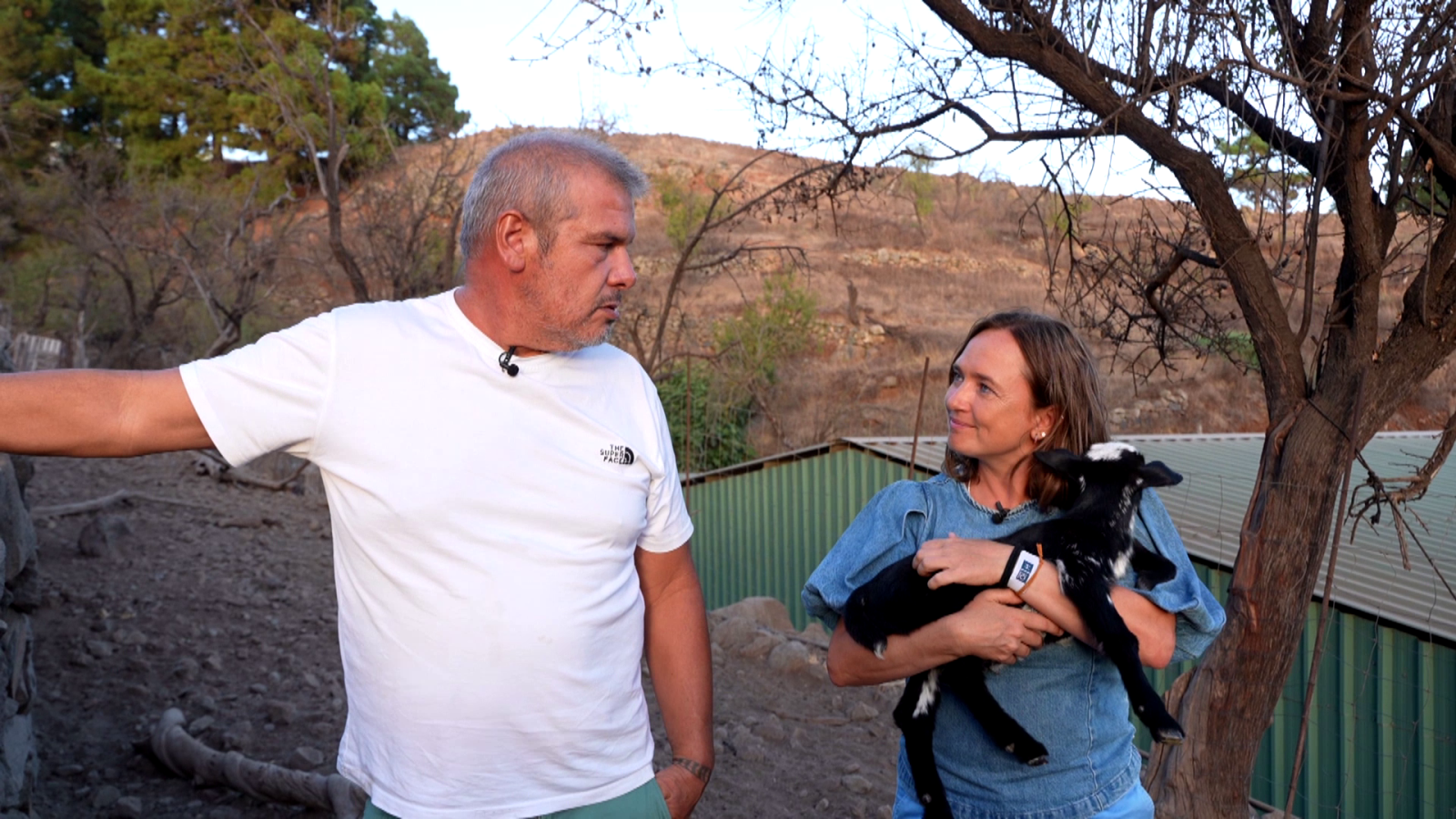 Reportero de "Aquí la Tierra" entrevista a una mujer que sostiene un cordero en un paisaje árido. Se observa una estructura de chapa verde al fondo.